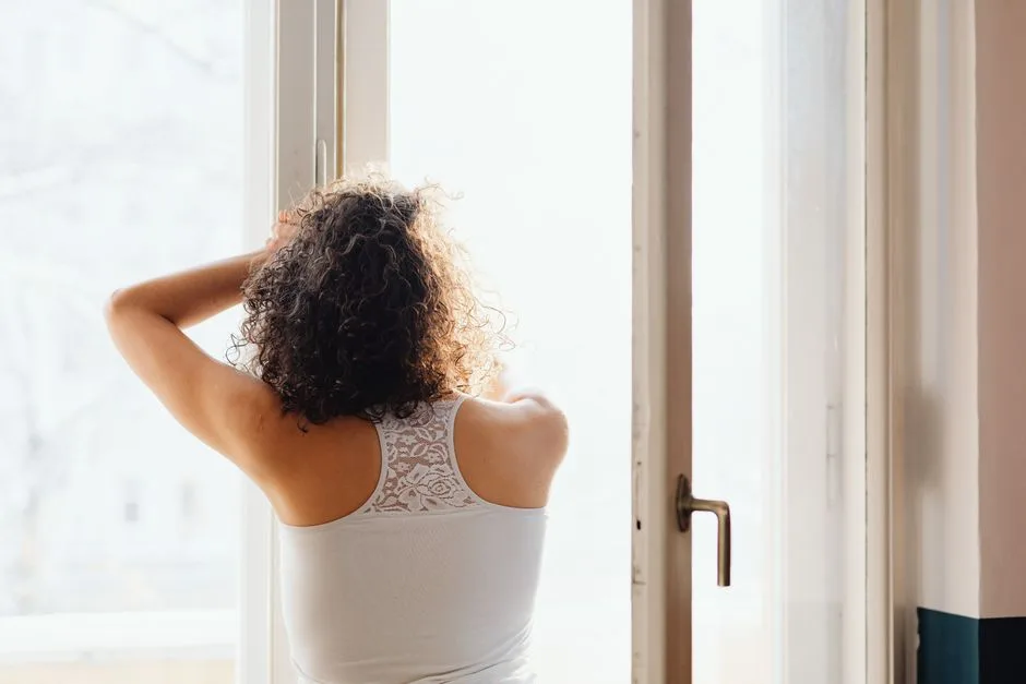 Femme de dos face à une grande fenêtre lumineuse, les mains dans les cheveux, qui expire calmement